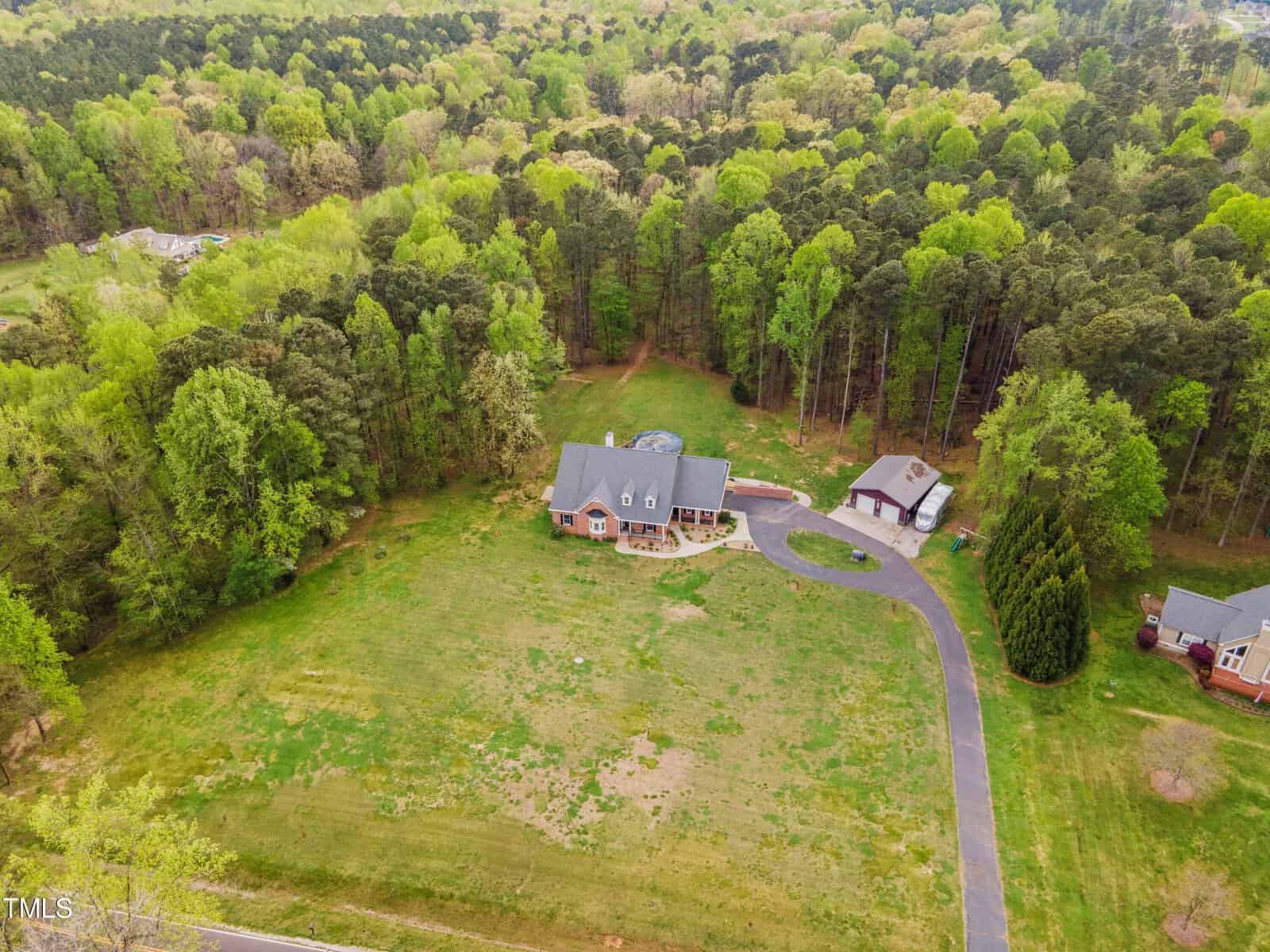 Aerial view of a rural property with a large green lawn, a paved driveway leading to a brick house with a gray roof, a circular pool, and a detached garage. Surrounded by dense trees and adjacent properties.