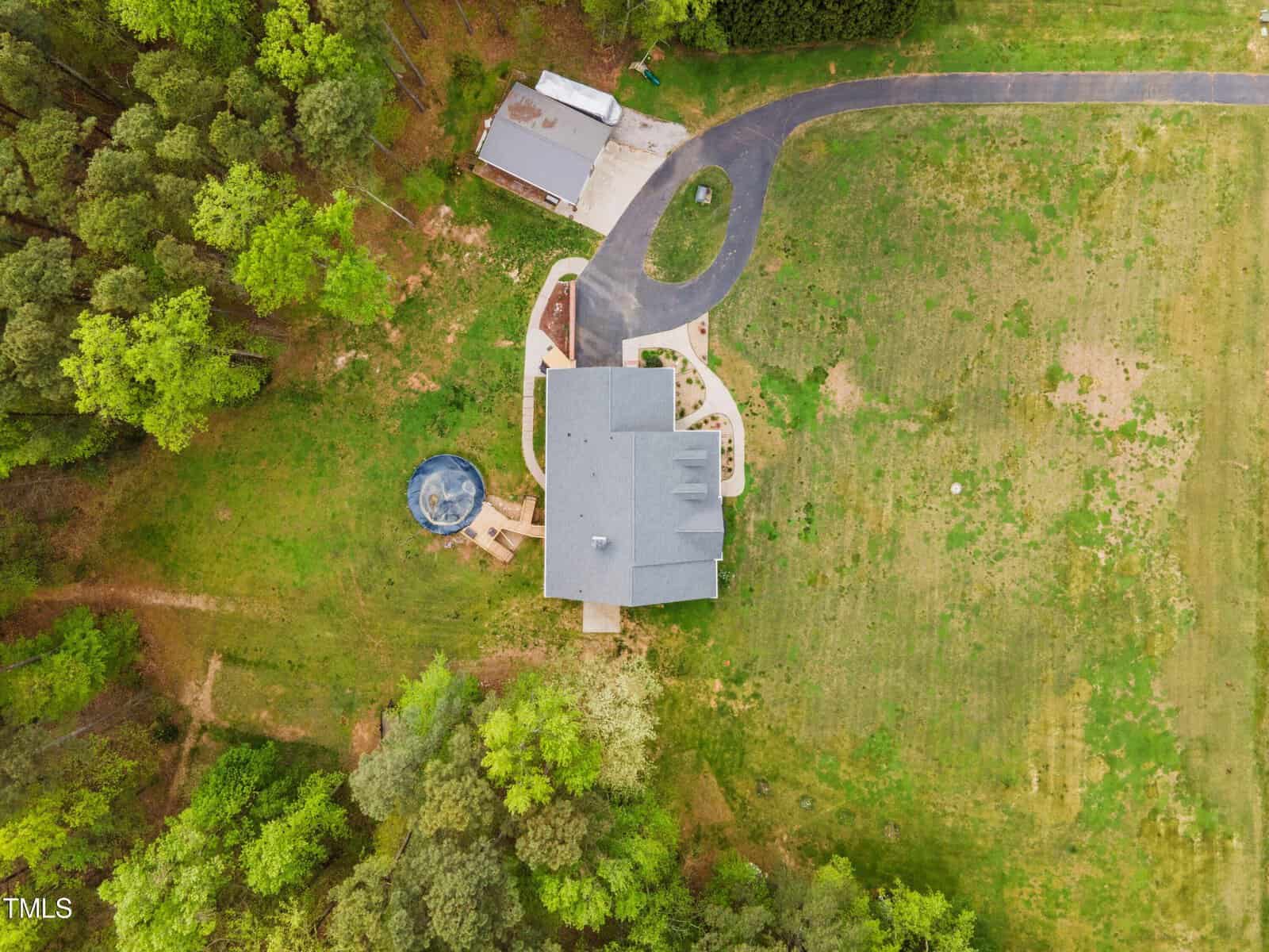 Aerial view of a house surrounded by green grass and trees. The house has a gray roof, a circular pool, and a winding driveway leading to a detached garage. The landscape includes manicured lawns and wooded areas.
