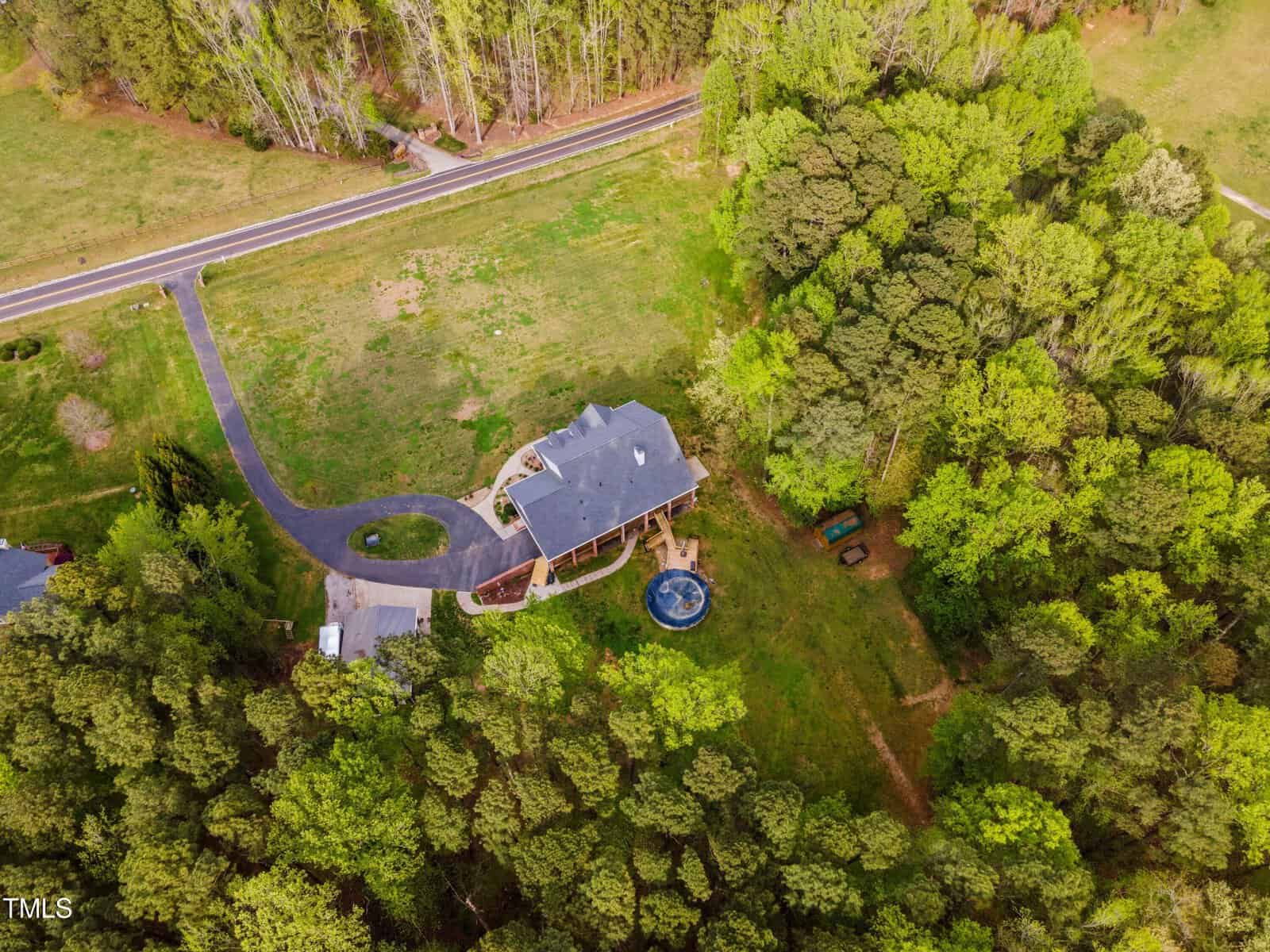 Aerial view of a house surrounded by dense green forest, with a curved driveway leading to it. There's a small pool in the backyard and a road visible nearby. The house has a spacious lawn and a wraparound deck.
