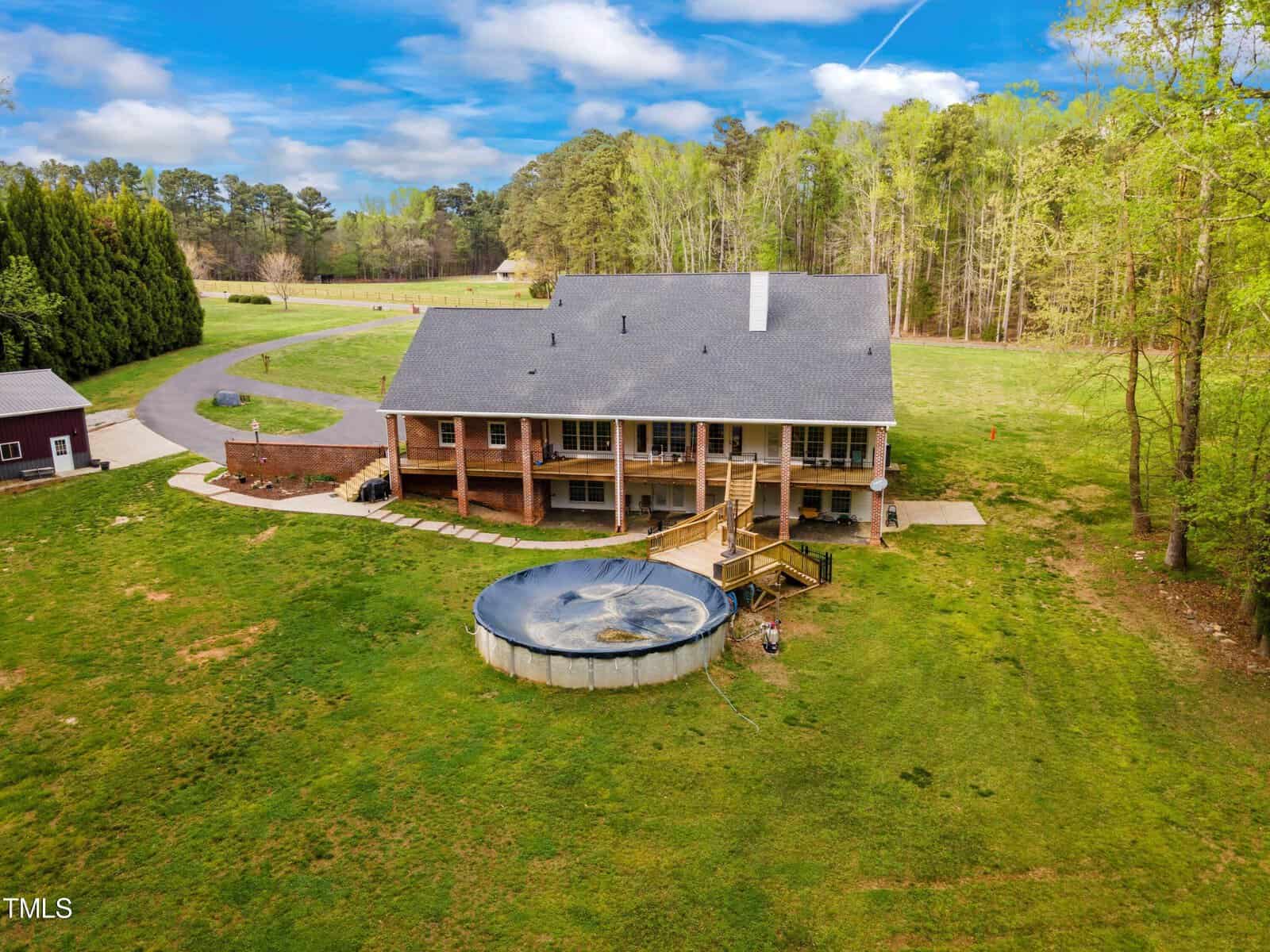 Aerial view of a large brick house with a spacious backyard, surrounded by trees. The backyard features a round above-ground pool and a wooden deck. A winding driveway leads to the front of the house. Sunny day with some clouds in the sky.
