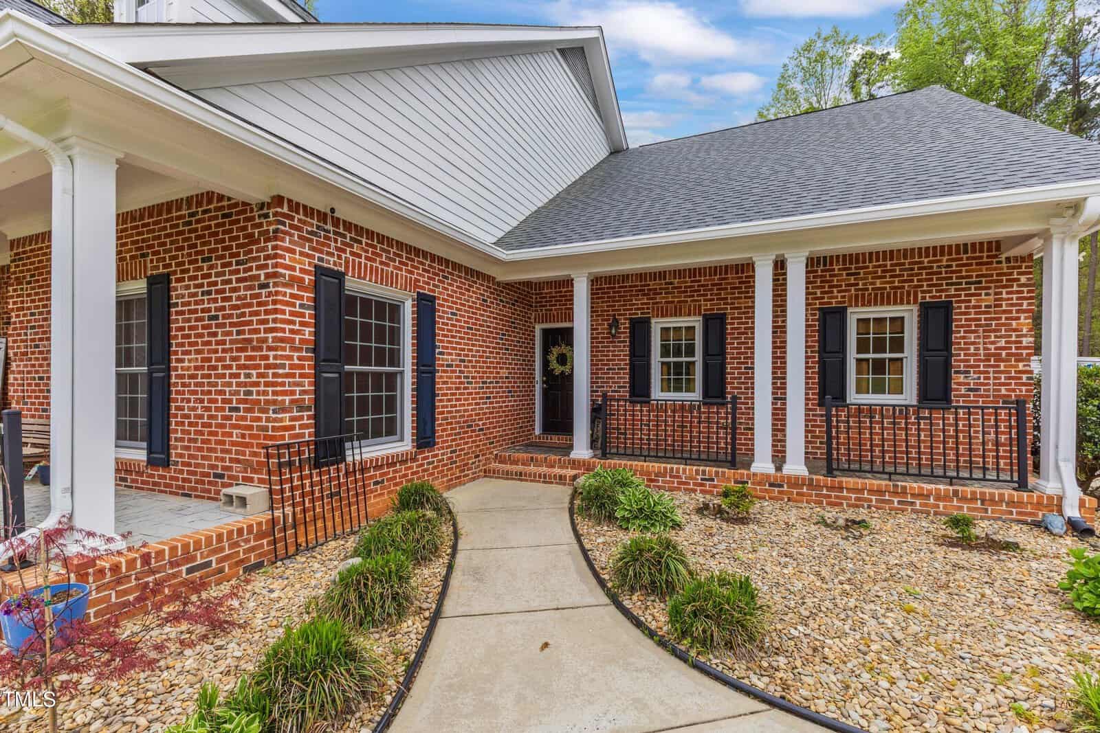 A brick house with white pillars and dark shutters, featuring a covered porch. The walkway is lined with plants and rocks. A wreath hangs on the front door, and the roof is dark gray. A bright, cloudy sky is in the background.