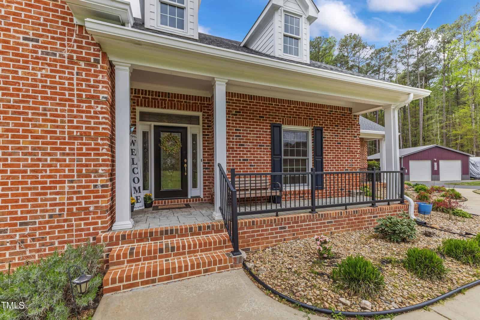 A brick house with a welcoming front porch featuring white columns, a black front door with a "Welcome" sign, and a black railing. Steps lead up to the porch, and there is tidy landscaping with shrubs in front. A detached garage is visible in the background.