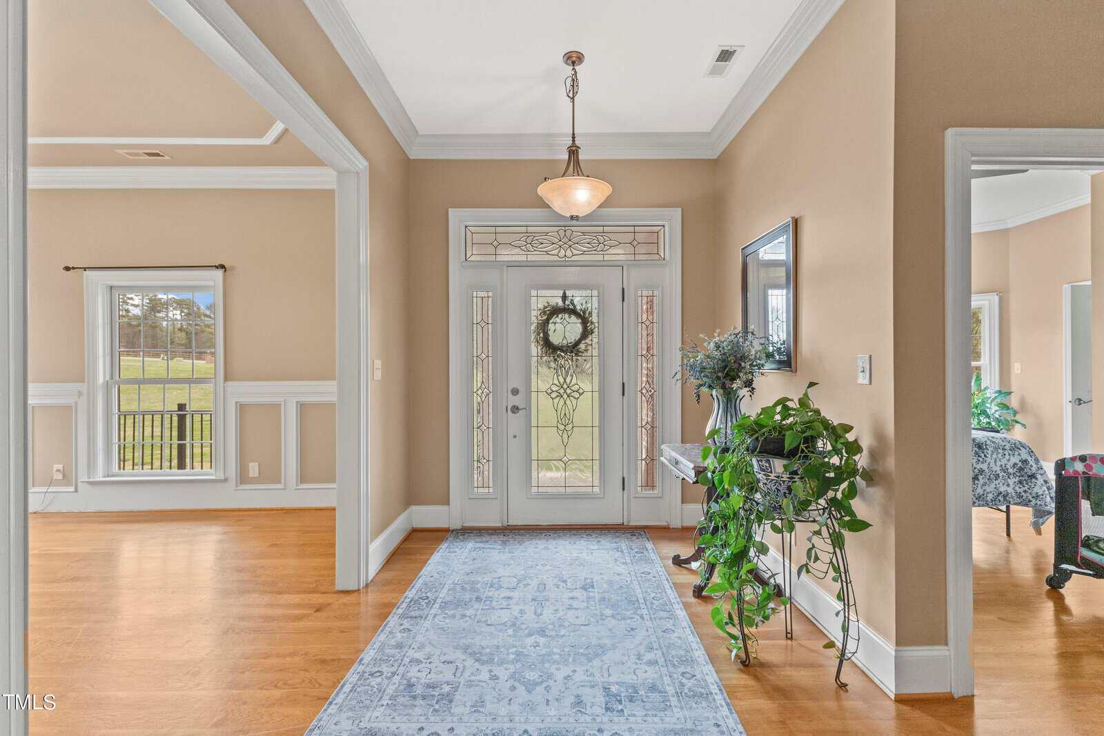 A brightly lit hallway with hardwood floors and beige walls. There's a blue rug leading to a glass front door adorned with a wreath. Potted plants are on either side, and a ceiling light fixture hangs above. A window shows a grassy outdoor view.