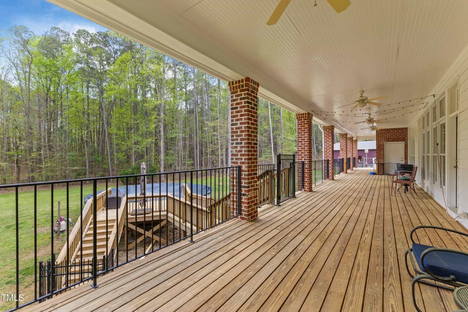 A spacious, covered wooden deck with ceiling fans overlooks a grassy backyard and a pool surrounded by a deck. Brick pillars support the roof, and string lights add charm. A few chairs are arranged for seating, creating a serene outdoor space.