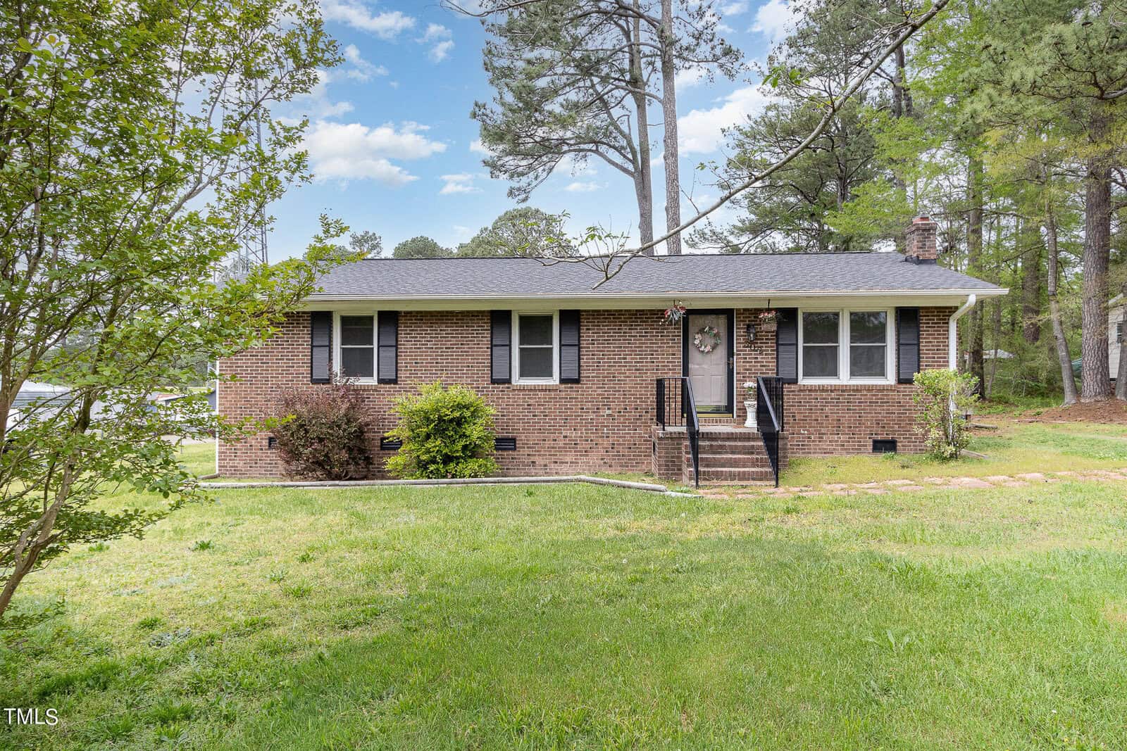 A charming single-story brick house at 302 W F Street in Butner boasts a black roof, surrounded by lush green grass and towering trees under a blue, cloud-kissed sky. The home features several windows with elegant black shutters and a welcoming door with steps leading up to it.