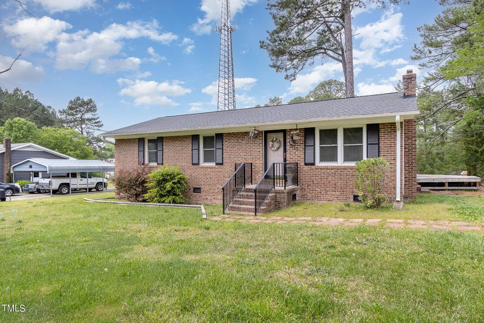 A brick single-story house with a gray roof and white trim set in a grassy yard. Steps lead up to the front entrance. Tall trees surround the property, and a tall radio tower is visible in the background. Blue sky with scattered clouds.