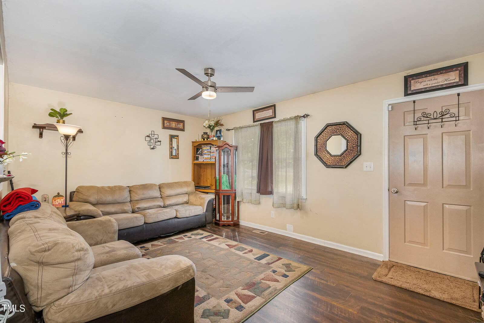 A cozy living room with two beige sofas, a wooden cabinet, and wall decorations. There's a ceiling fan, a window with sheer curtains, and a light-colored door. A patterned rug is on the dark hardwood floor.