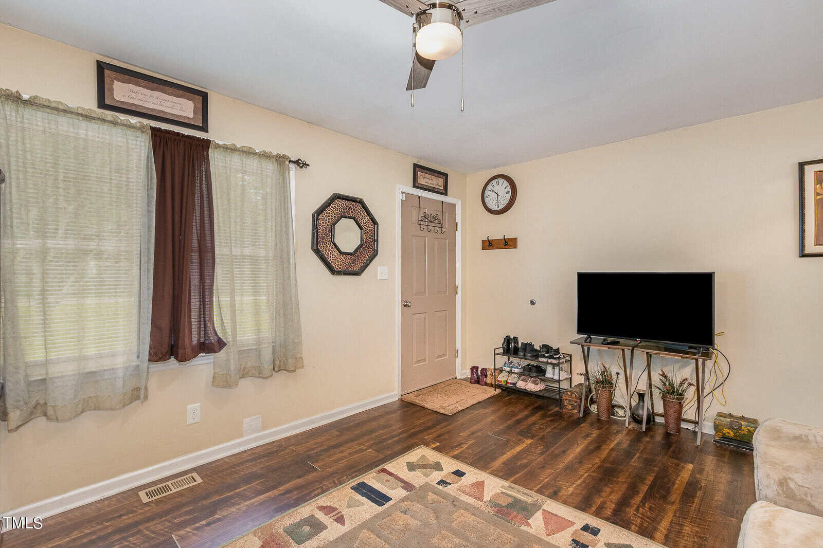 A cozy living room features wooden floors, a TV on a stand, a ceiling fan, and a door with a wall clock above. Decor includes framed art, a patterned rug, and indoor plants. A window with curtains allows natural light to filter into the space.