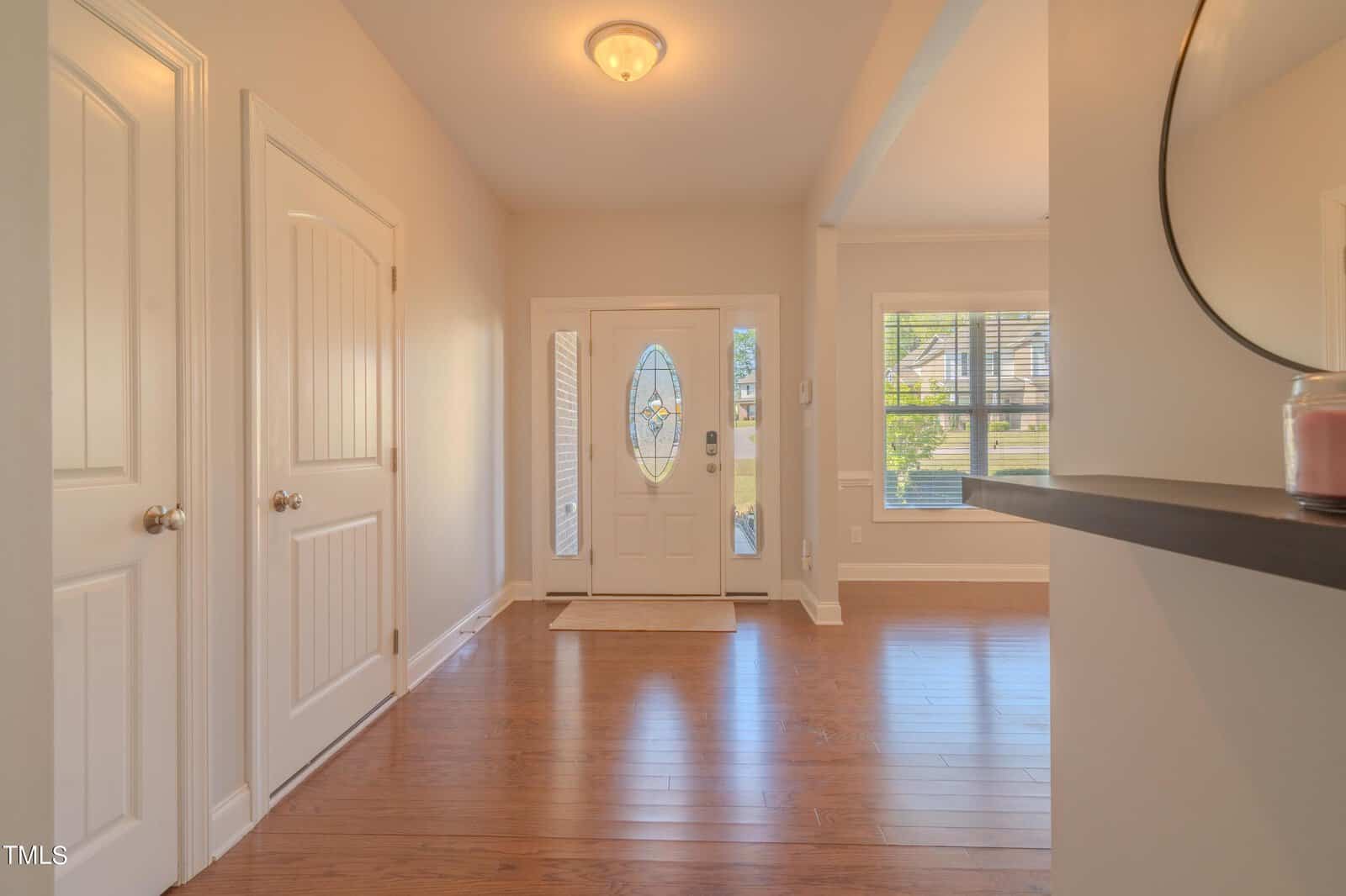 A bright entrance hallway with wooden floors, white walls, a central light fixture, and a glass-paneled white front door. There are two closed doors on the left and a rectangular mirror on the right wall.