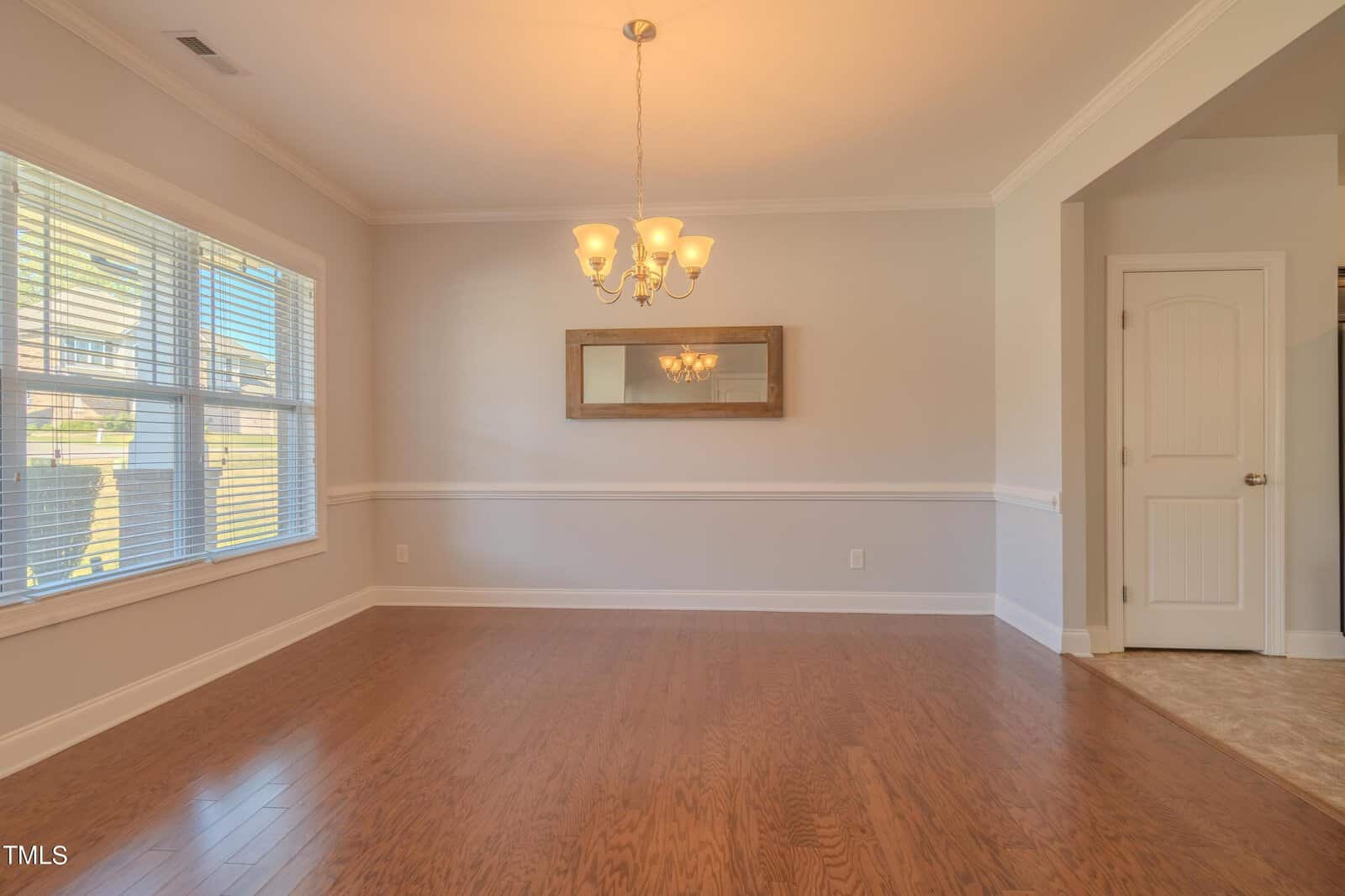 A bright, empty room with hardwood floors, a chandelier, and a large window. There's a rectangular mirror on the wall and a doorway leading to another area. Natural light fills the space, highlighting the neutral wall colors.