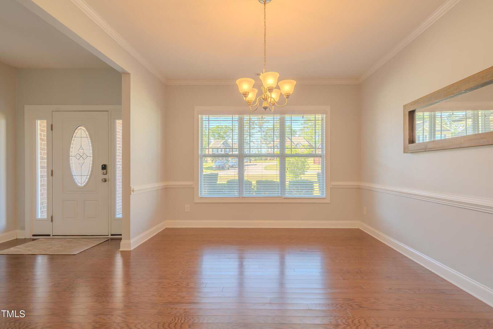 A bright, empty room features wooden floors and a large window with white blinds, letting in natural light. A chandelier hangs from the ceiling. There's a white front door to the left and a mirror on the right wall.