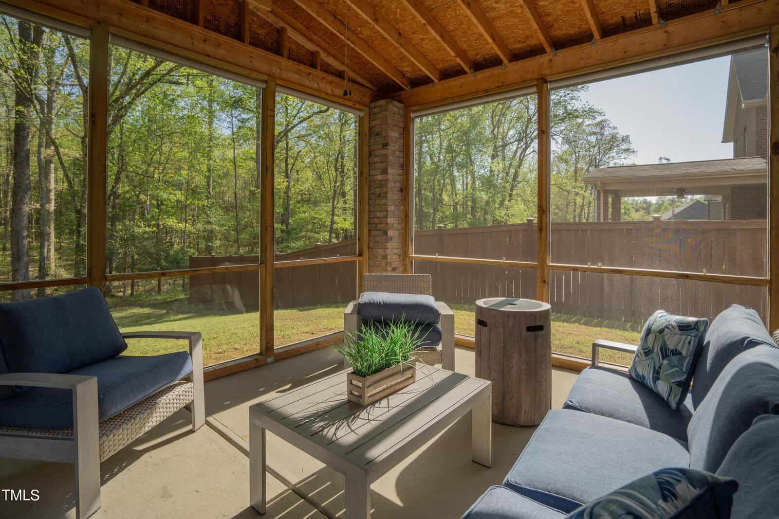 A cozy outdoor screened porch with comfortable blue cushioned seating set around a white coffee table. A potted plant sits on the table. The porch overlooks a green yard through large screened windows, framed by wooden beams.