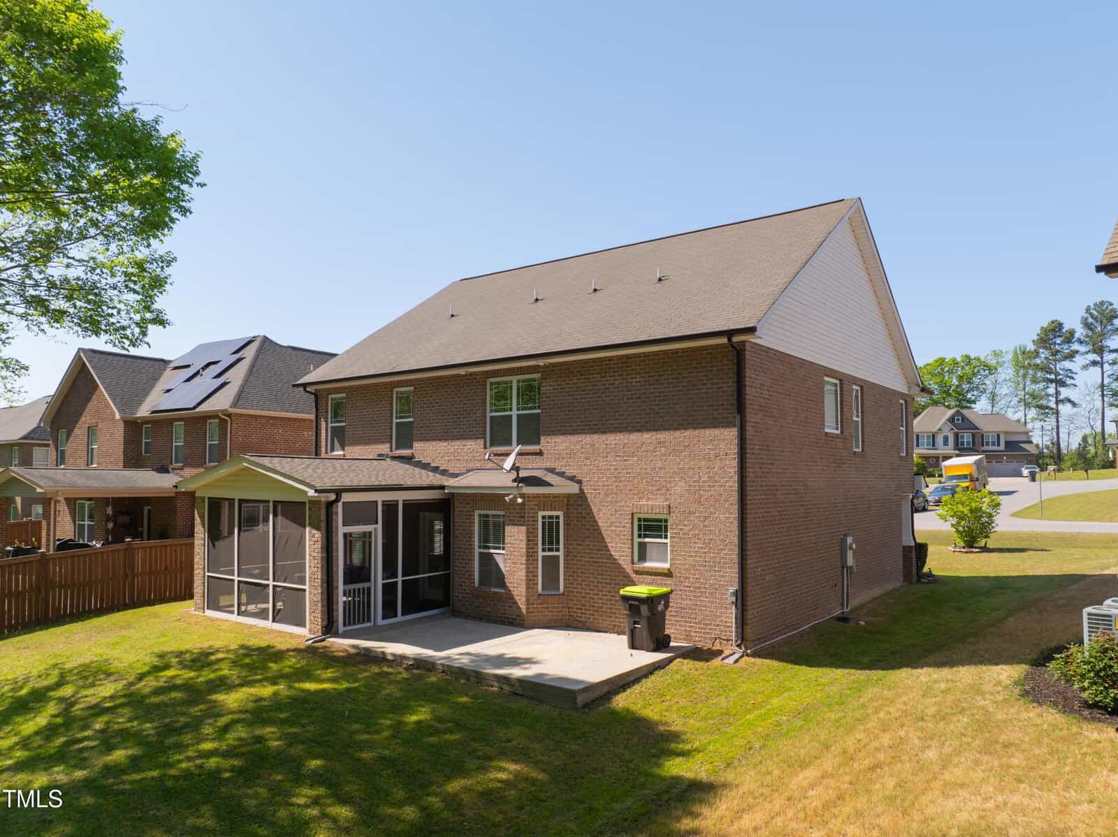A two-story brick house with a screened porch in a suburban neighborhood. The backyard features a grassy lawn and a concrete patio. Adjacent houses and a clear blue sky are visible in the background.