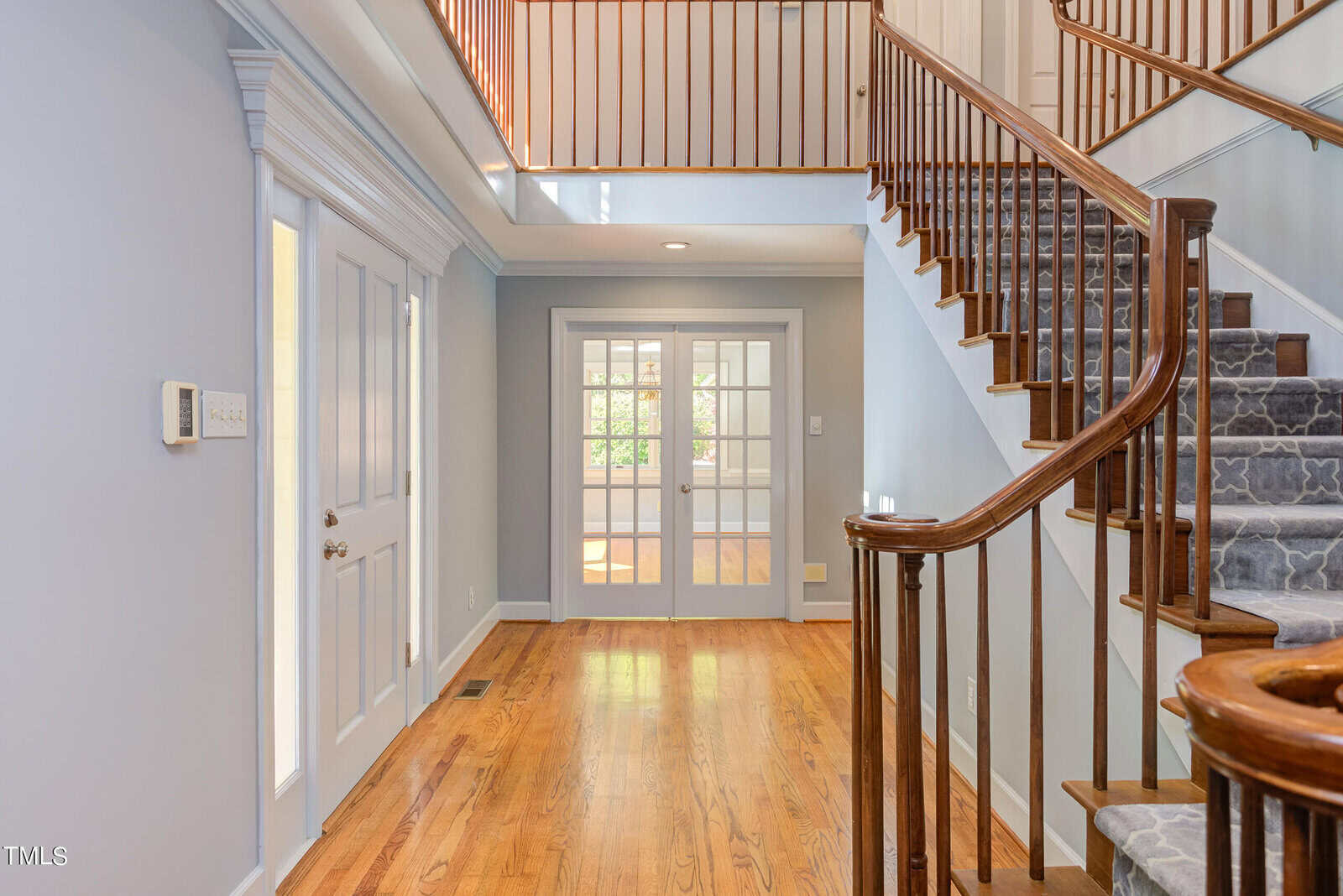 A bright hallway with wooden flooring and a curved staircase. Light blue walls and white trim enhance the space, leading to double French doors at the end. Upper balcony railing matches the stairs. Natural light streams in from the doors.