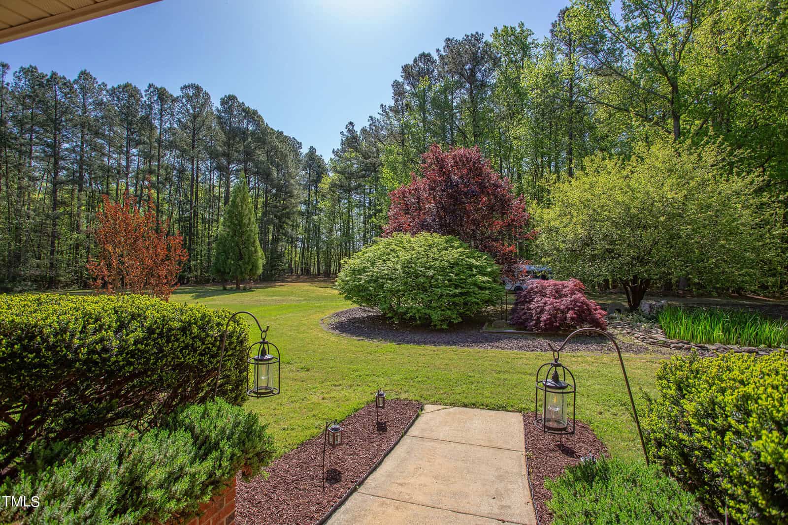 A scenic view of a garden with a paved path leading through lush green grass, surrounded by trimmed hedges. Colorful trees, including red and green foliage, border the yard. Two lanterns stand along the path, and tall pine trees line the background.