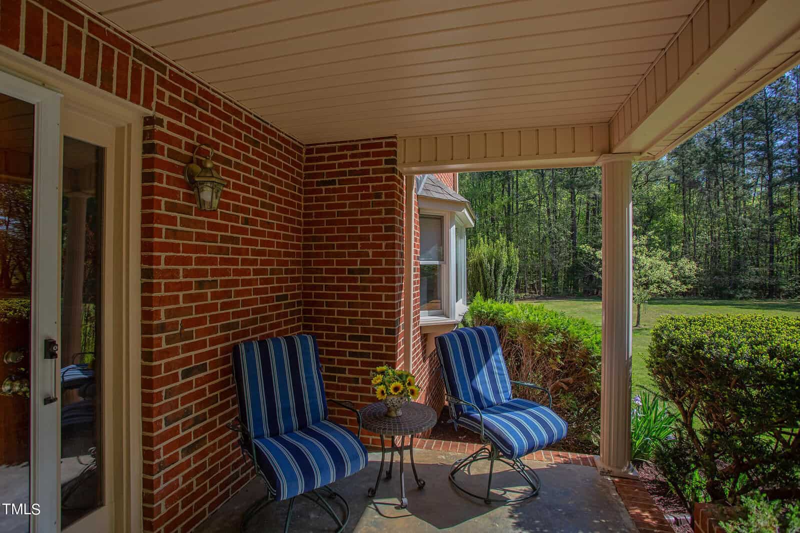 A cozy brick patio with two blue-striped cushioned chairs and a small round table holding a vase of sunflowers. The area is surrounded by lush greenery and bushes, with a view of a wooded area in the background.