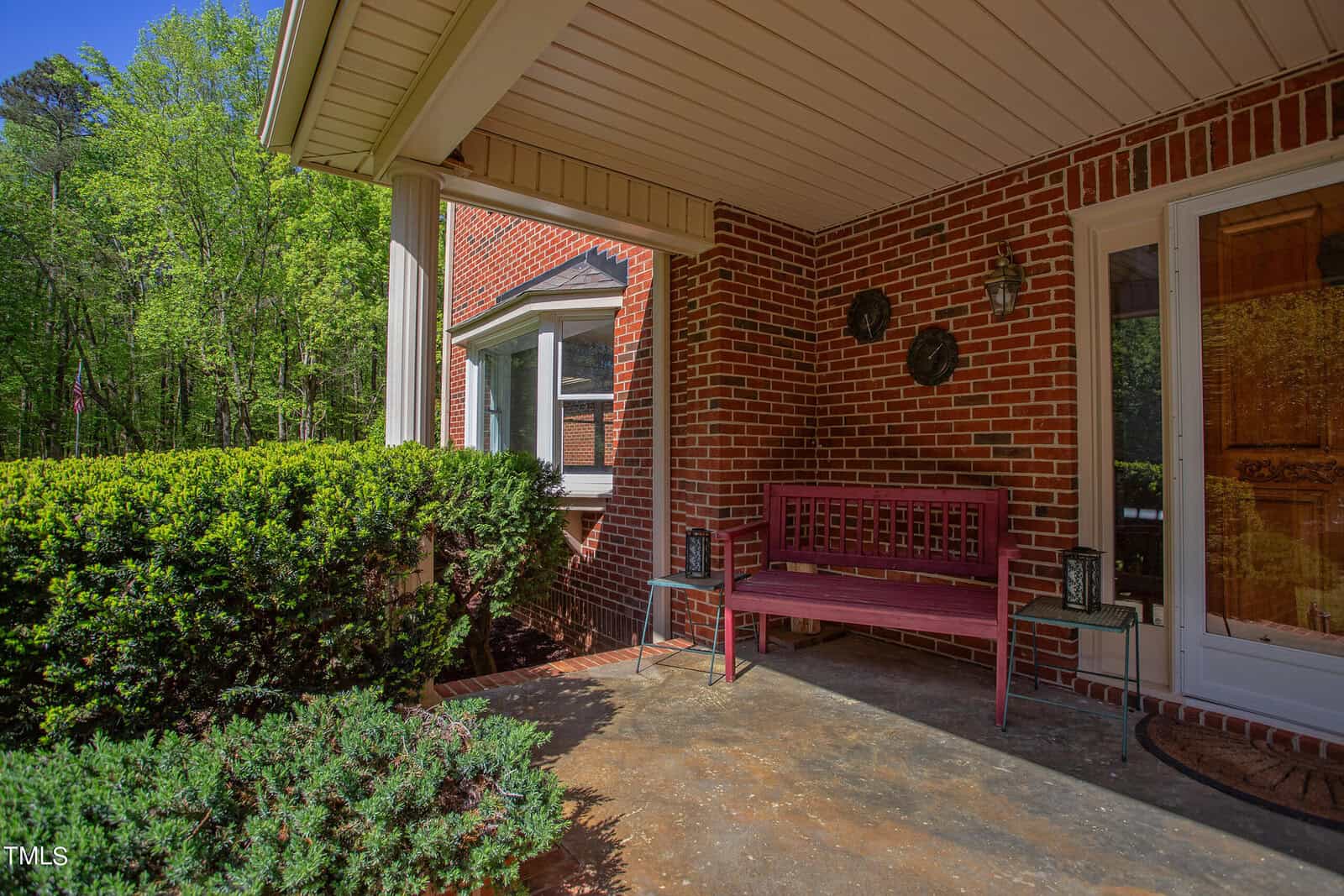 A red brick house porch with a wooden bench, decorative wall ornaments, and neatly trimmed bushes. The porch is shaded, offering a view of green trees in the background under a bright blue sky.