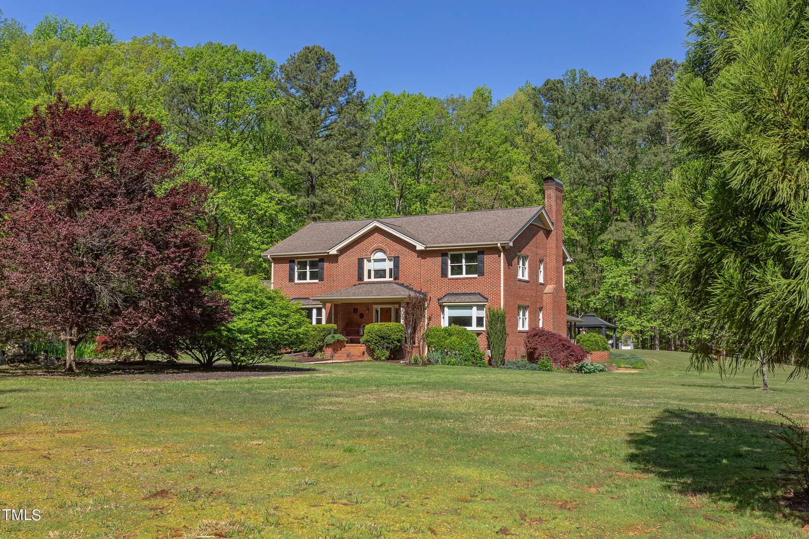 A large, two-story brick house is surrounded by lush greenery and trees. The front yard is spacious with well-maintained grass and a variety of shrubs, including a red-leaved tree on the left. A clear blue sky is in the background.