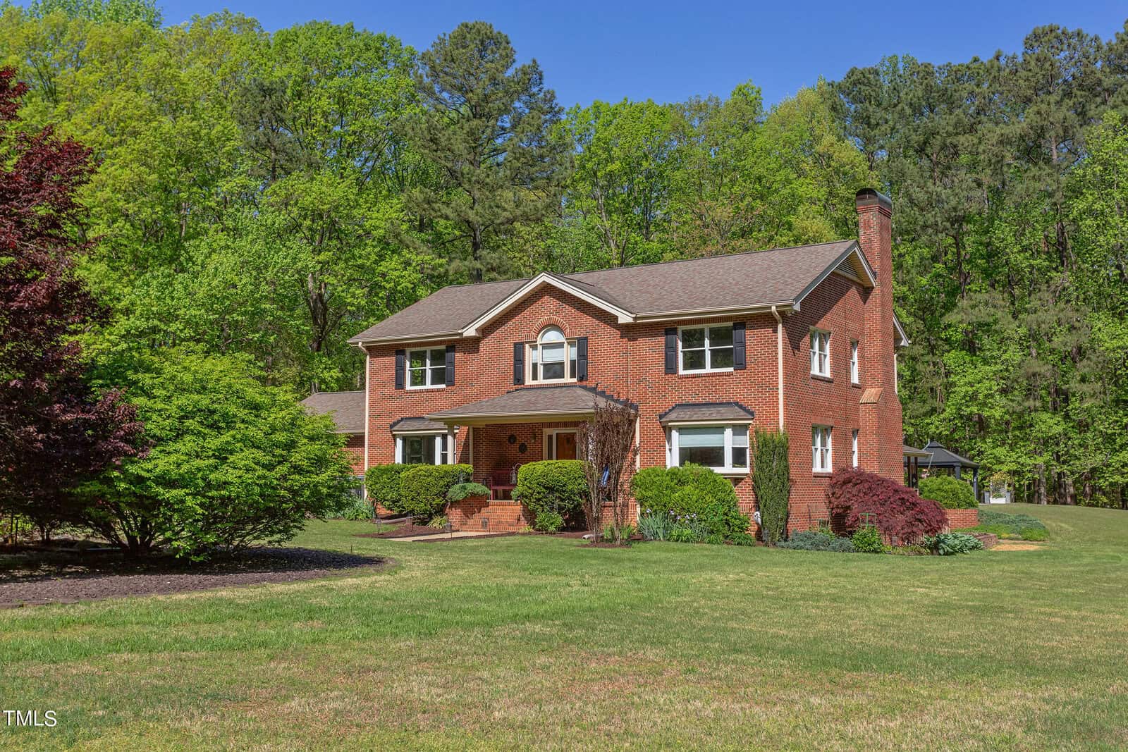 A large two-story red brick house with a brown roof set in a lush green landscape. Manicured lawns and vibrant bushes surround the home, with tall trees in the background under a clear blue sky.