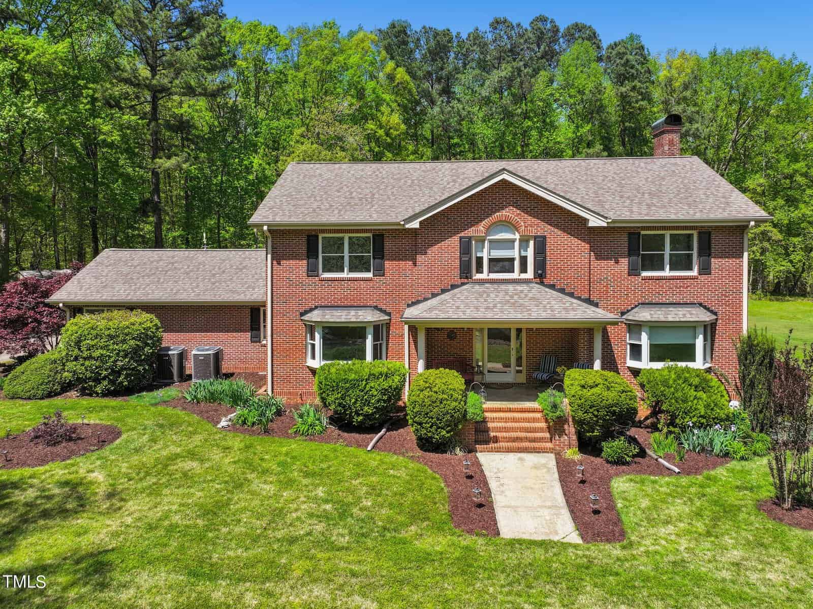 The charming two-story red brick house at 4057 Chewning Road, Oxford boasts a neatly manicured lawn and hedges. With two windows on each side of the central entrance and a chimney on the right, it sits against a backdrop of lush green trees under a clear blue sky.
