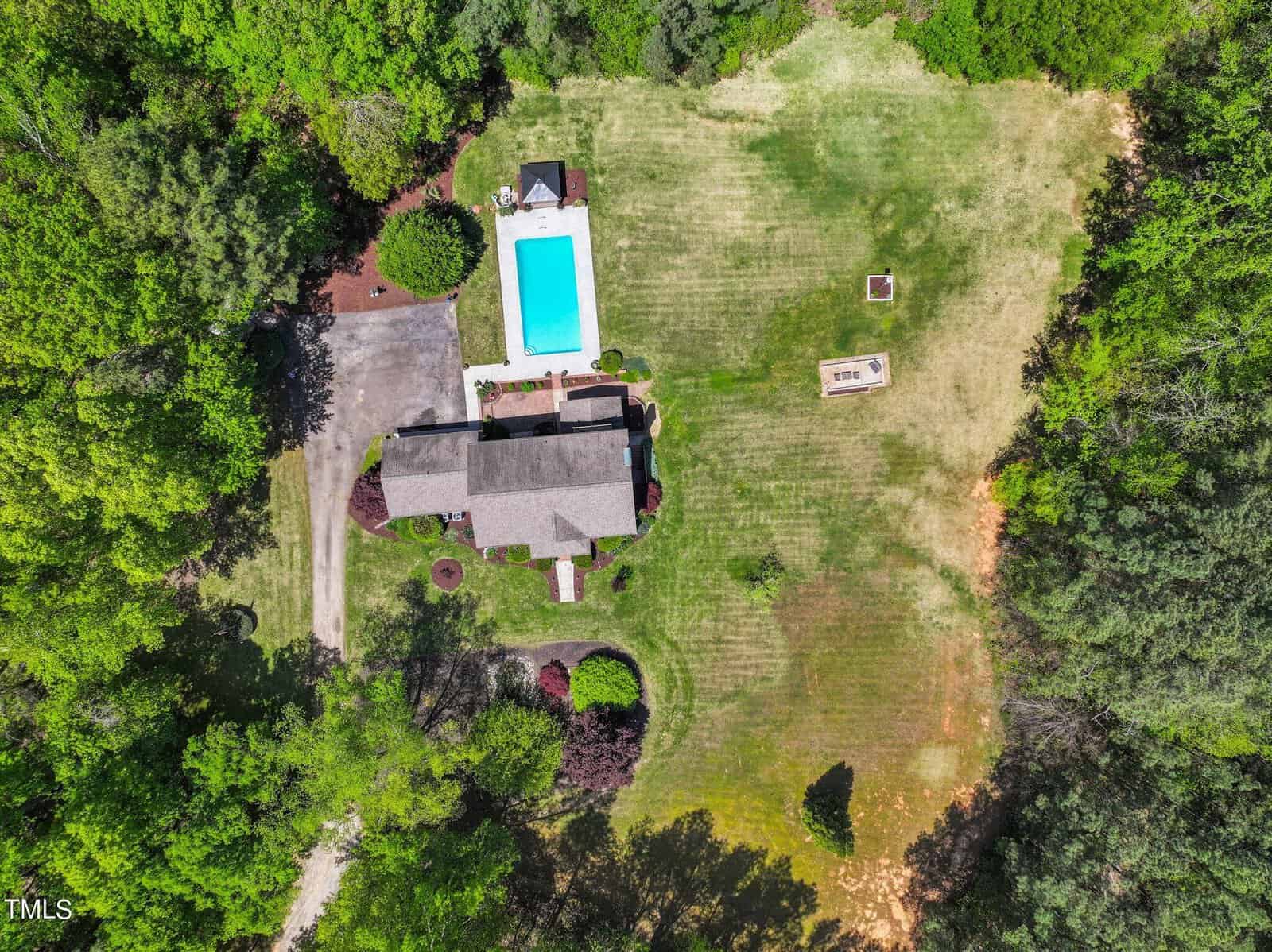 Aerial view of a house surrounded by trees and grassy fields. There is a rectangular swimming pool with a small patio area beside it. A long driveway leads to the house, and there's a separate fire pit area on the lawn.