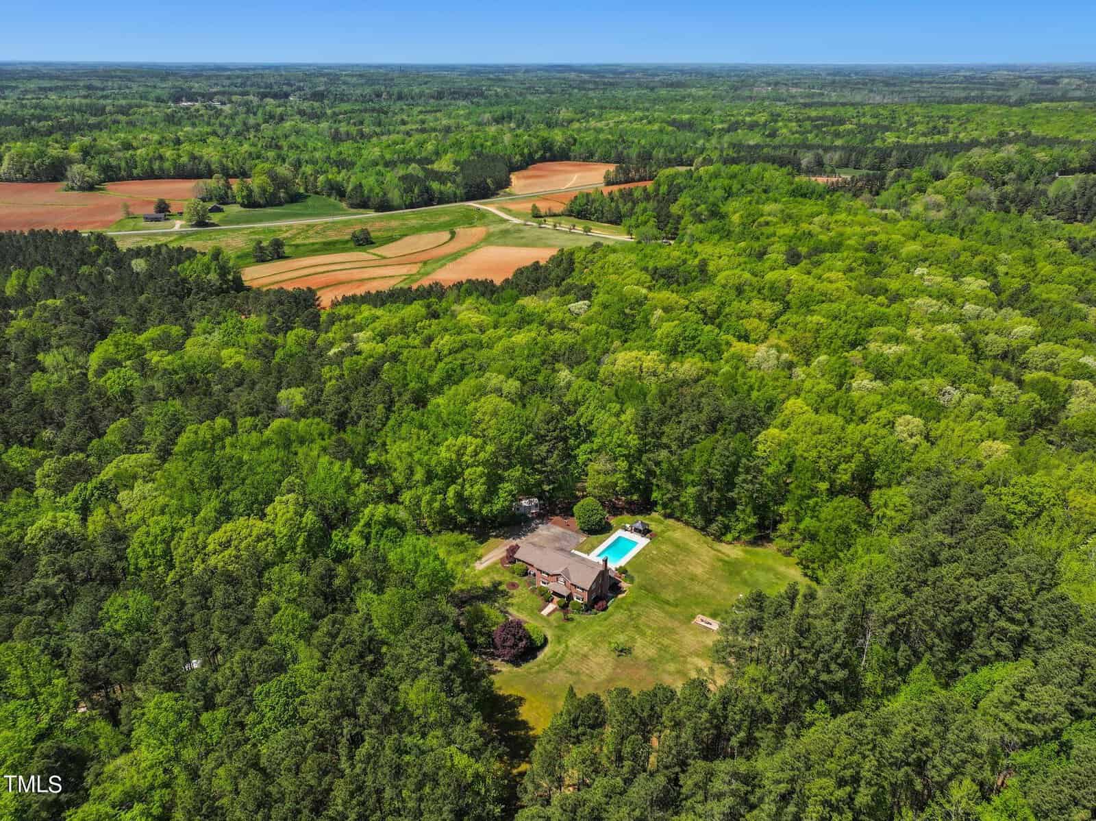 Aerial view of a secluded house with a pool surrounded by dense green forest and fields. The landscape features lush vegetation and open farmland under a clear blue sky.