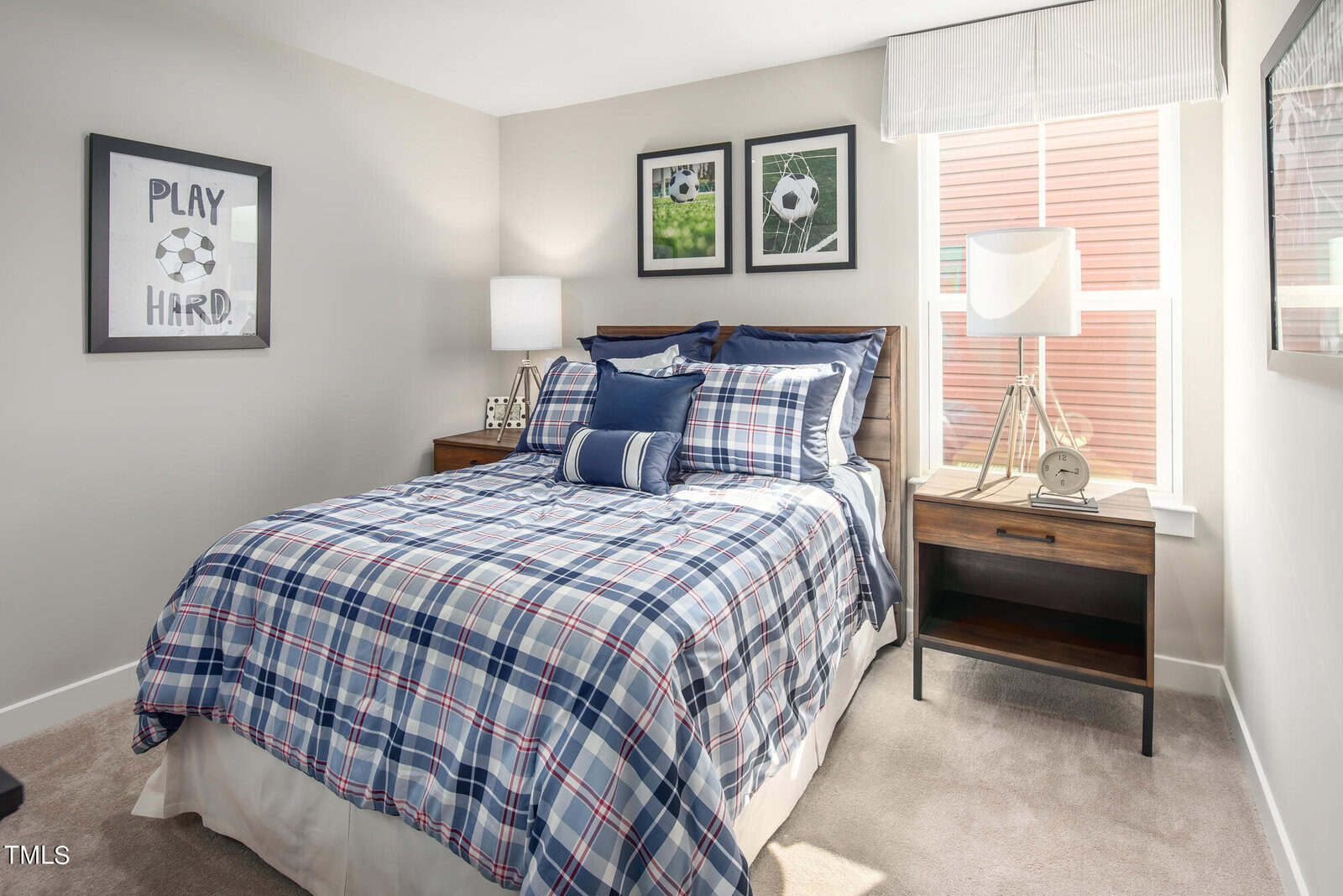 Bright bedroom with a neatly made bed in blue plaid bedding, soccer-themed wall art, two nightstands with lamps, and large windows letting in natural light.