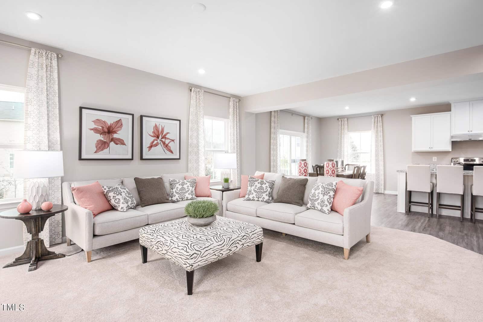 Bright, modern living room with two beige sofas, patterned ottoman, pink and gray accent pillows, floral artwork, tall windows with light curtains, and an open view into a white kitchen and dining area.
