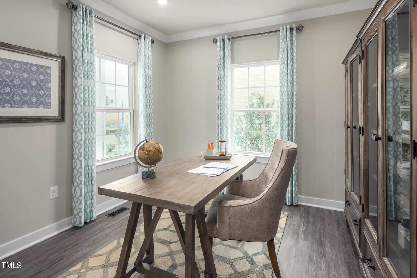 A bright home office with a wooden desk, padded chair, globe, and papers. Two large windows with patterned curtains let in natural light. There's a glass-front cabinet and geometric rug on the floor.