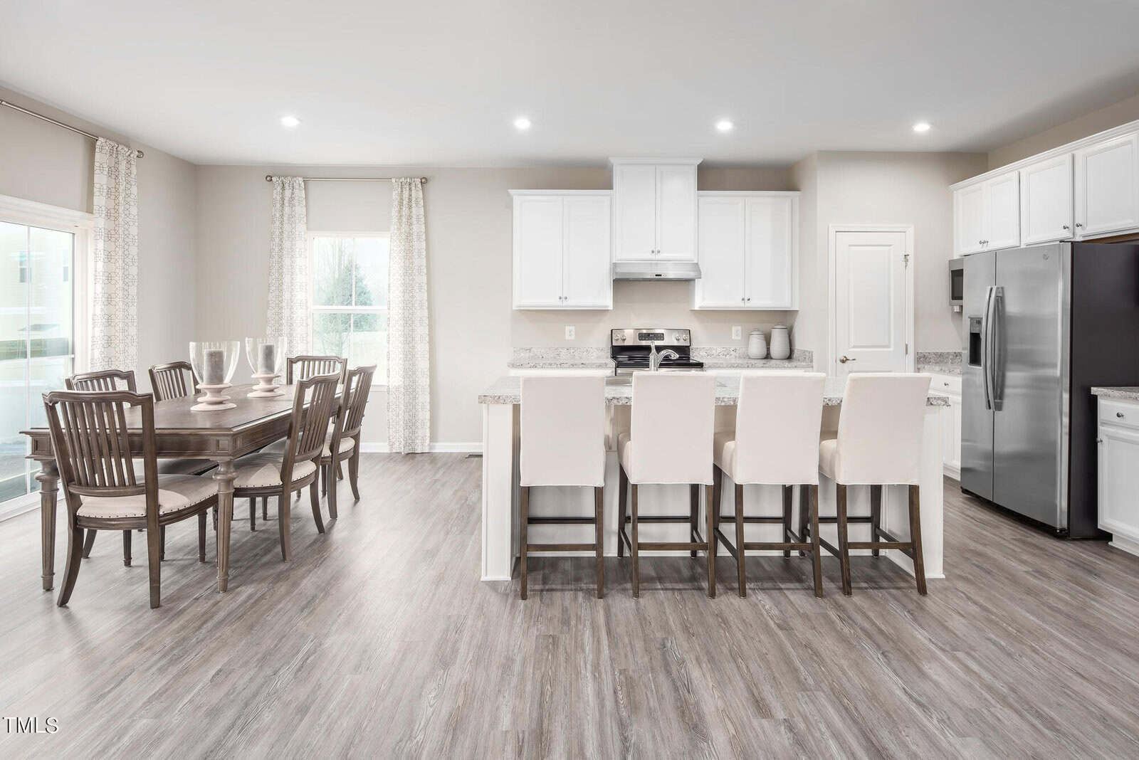 Modern kitchen and dining area with white cabinets, stainless steel appliances, a marble countertop island with four beige barstools, and a wooden dining table with six chairs on a light wood floor.