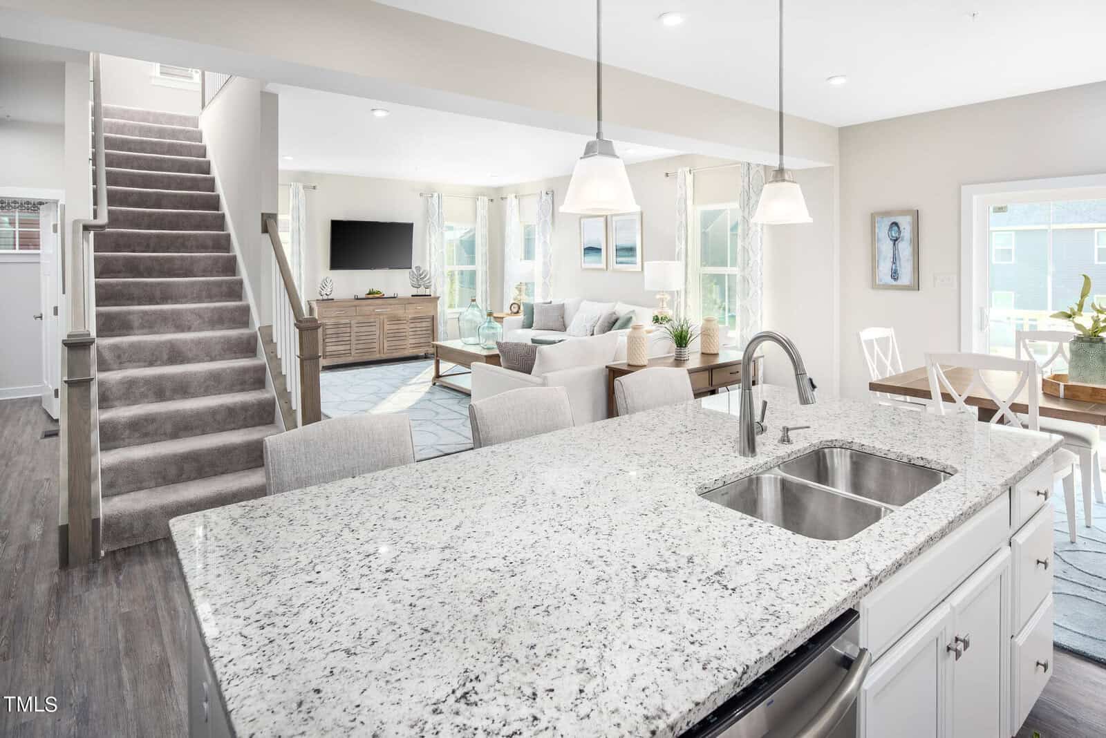 Bright, modern open-concept living space with a granite kitchen island, white cabinets, dining area, living room with a sofa and TV, large windows, and a carpeted staircase leading upstairs.