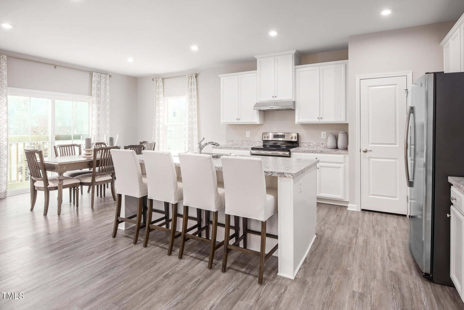 Modern kitchen with white cabinets, stainless steel appliances, a marble island with four white barstools, and a dining area with a wooden table and chairs by large windows with curtains. Light wood flooring throughout.