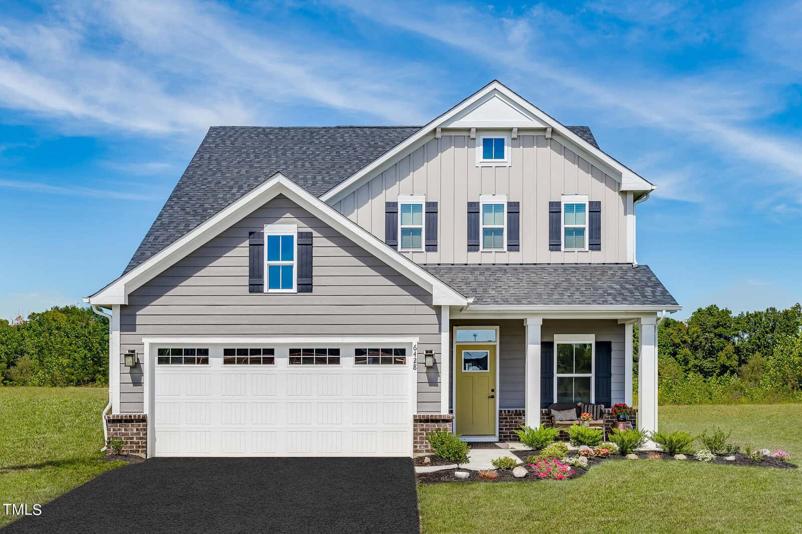 A two-story modern suburban house with gray siding, white trim, and a double garage, situated on a green lawn with a small landscaped garden and a clear blue sky in the background.