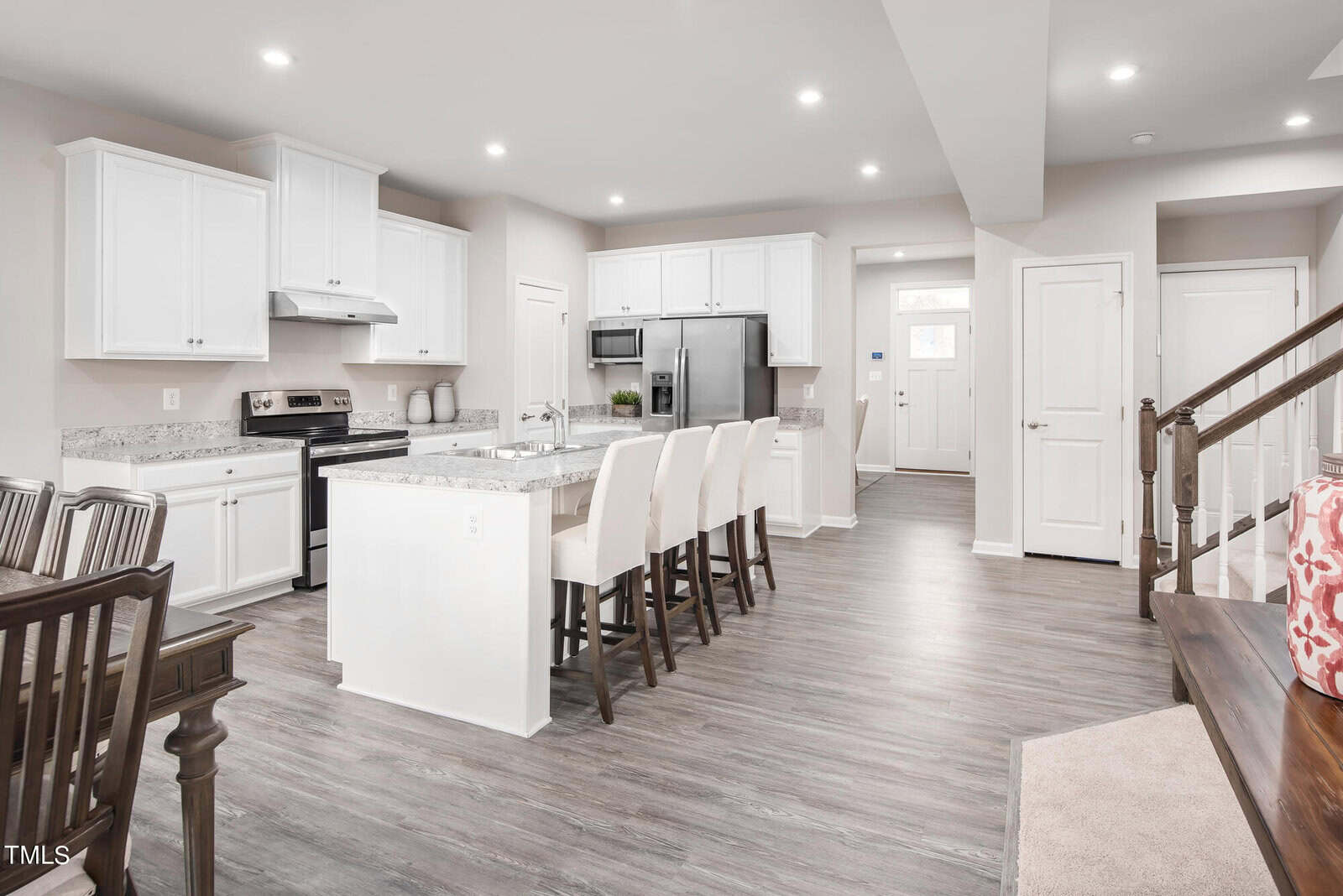 Modern kitchen with white cabinets, stainless steel appliances, and an island with four white barstools. Open floor plan with a dining area, wood flooring, and a staircase on the right. Bright and spacious atmosphere.