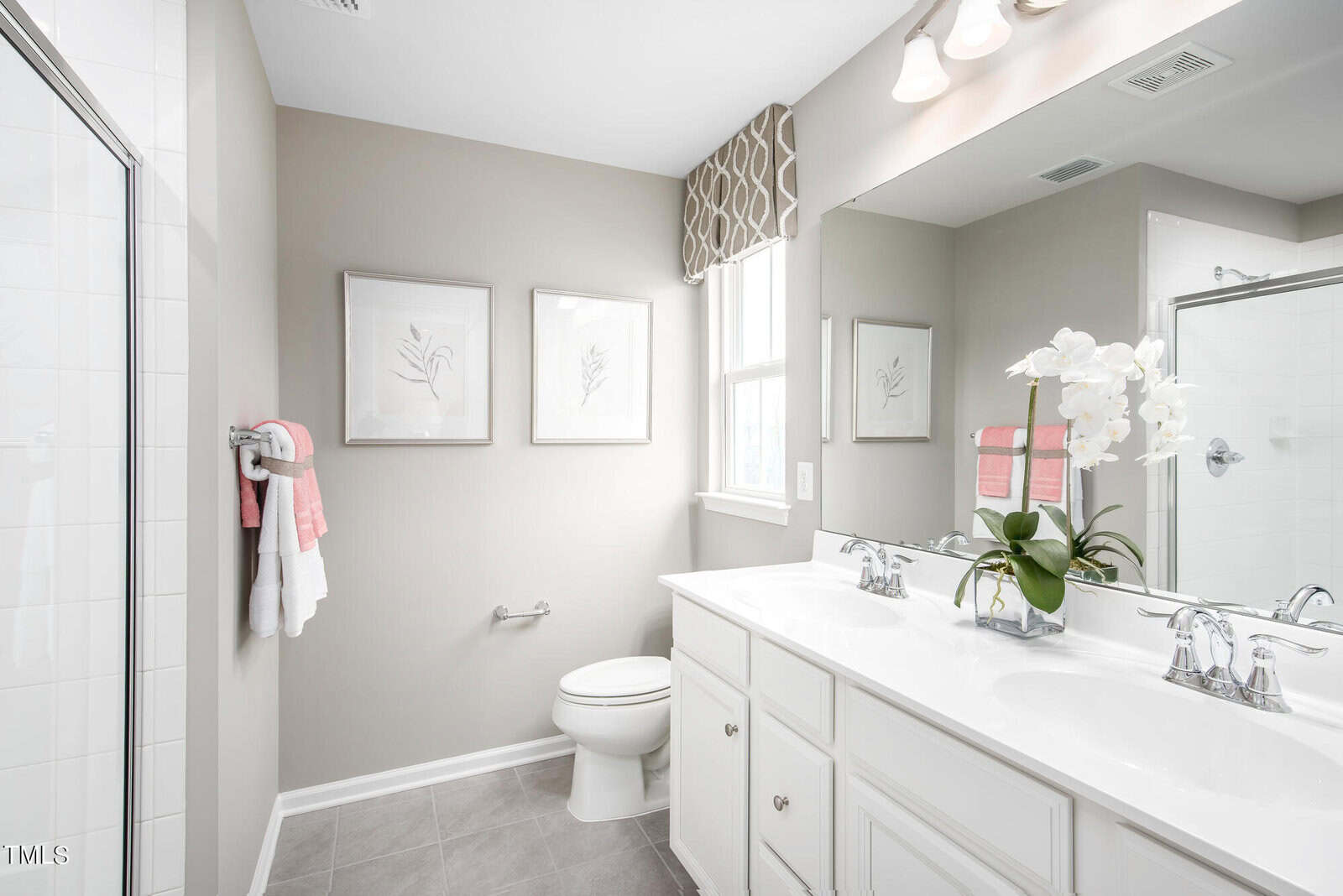 Bright, modern bathroom with white cabinets, double sinks, a large mirror, gray floor tiles, and beige walls. There are two framed pictures, a window, a toilet, and a shower. A white orchid decorates the vanity.
