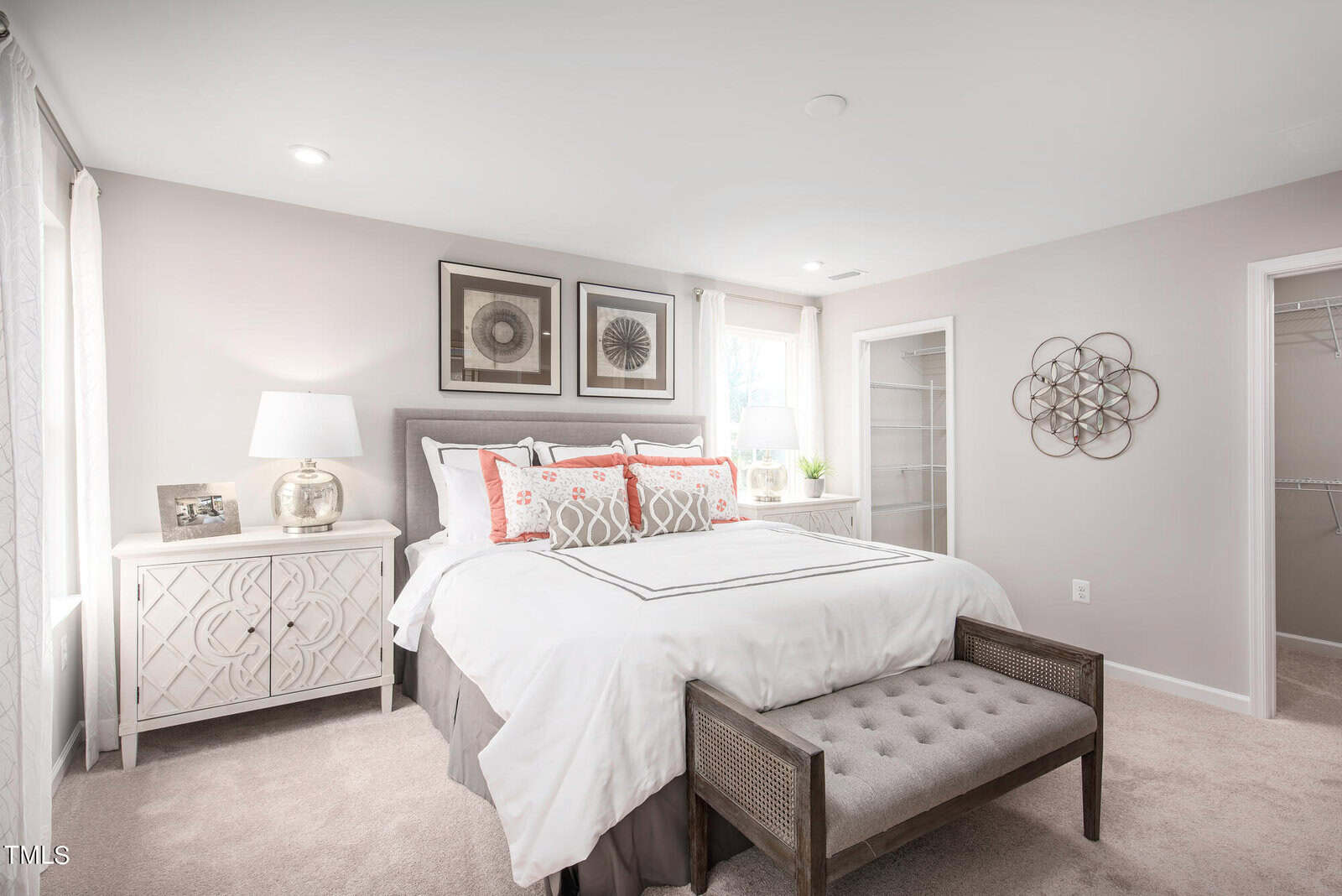 Bright bedroom with a neatly made bed, gray bench at the foot, white nightstand with lamp and photo, wall art above the bed, and walk-in closets with open doors. Light carpets and neutral walls complete the room.