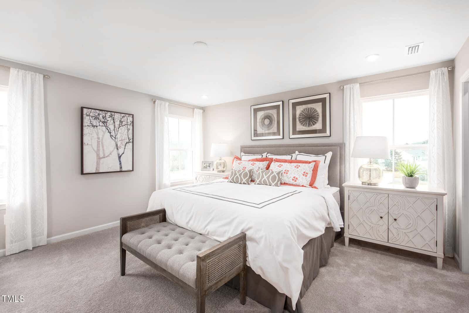A bright, modern bedroom with a large bed featuring white bedding and red accent pillows, two nightstands with lamps, a cushioned bench at the foot of the bed, and framed art on pale walls with two windows letting in natural light.