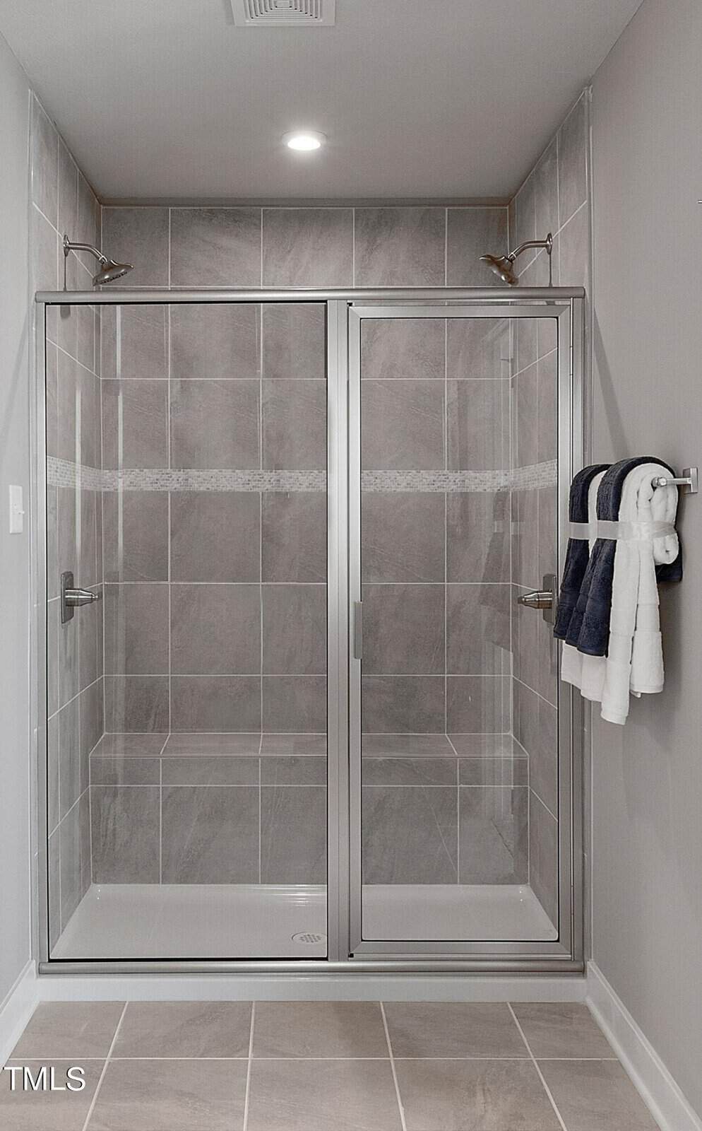 Modern double shower with gray tiles, glass doors, and built-in benches. A towel rack with neatly folded white and navy towels is mounted on the right wall. Ceiling light provides illumination.