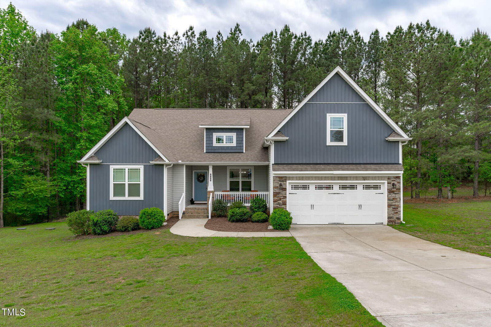 Located at 955 Weatherby Lane in Creedmoor, this gray two-story house features white trim and a gabled roof. It's set against dense trees and a vast lawn. The attached garage with white doors complements the concrete driveway leading to the street, while a cozy porch welcomes visitors near the entrance.