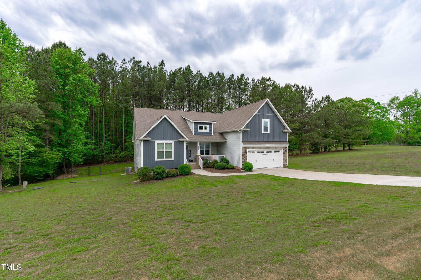 A modern gray house with a two-car garage situated in a spacious grassy yard. The home is bordered by a dense line of tall trees. A curved concrete driveway leads to the house, and the sky above is overcast.