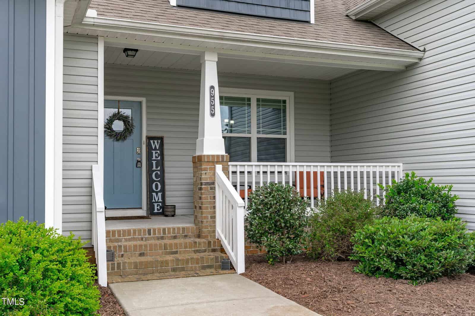 A house entrance with a blue door featuring a wreath. A black "Welcome" sign stands against a brick column. White railing surrounds a small porch with a window. Green shrubs line the walkway leading to the door.