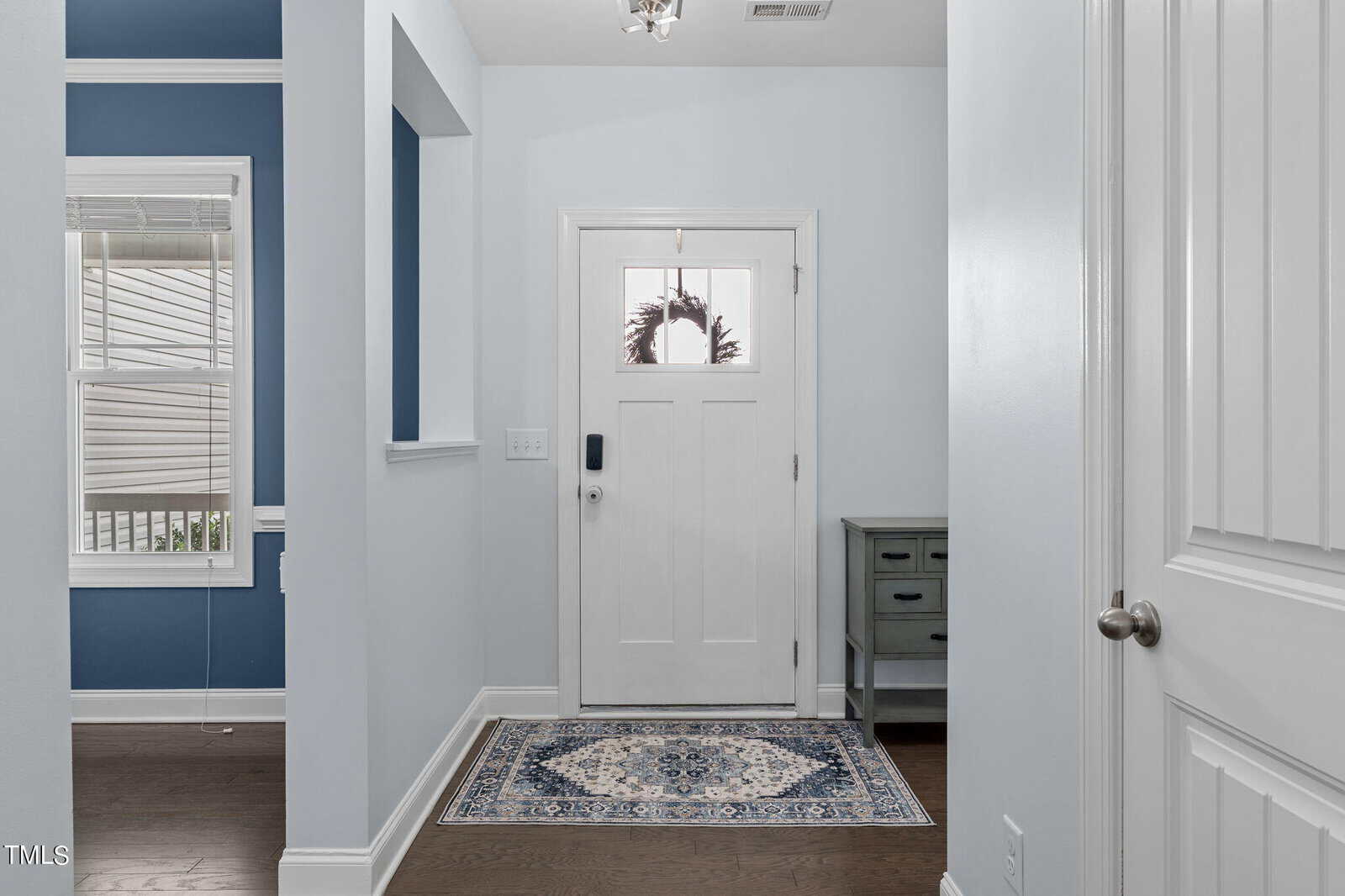 A modern, light-filled foyer with pale blue walls, a white front door with a window, and dark hardwood floors. A patterned rug lies on the floor, and a small cabinet sits near the door. Natural light illuminates the space.