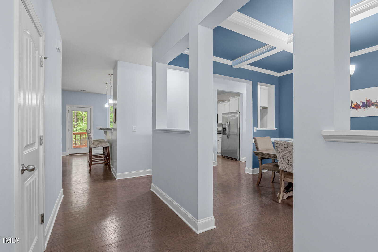 A modern home interior with light blue walls and white trim. The hallway leads to a dining area and kitchen with stainless steel appliances. Wooden flooring is throughout, and a door opens to a deck outside. Hanging lights illuminate the spaces.