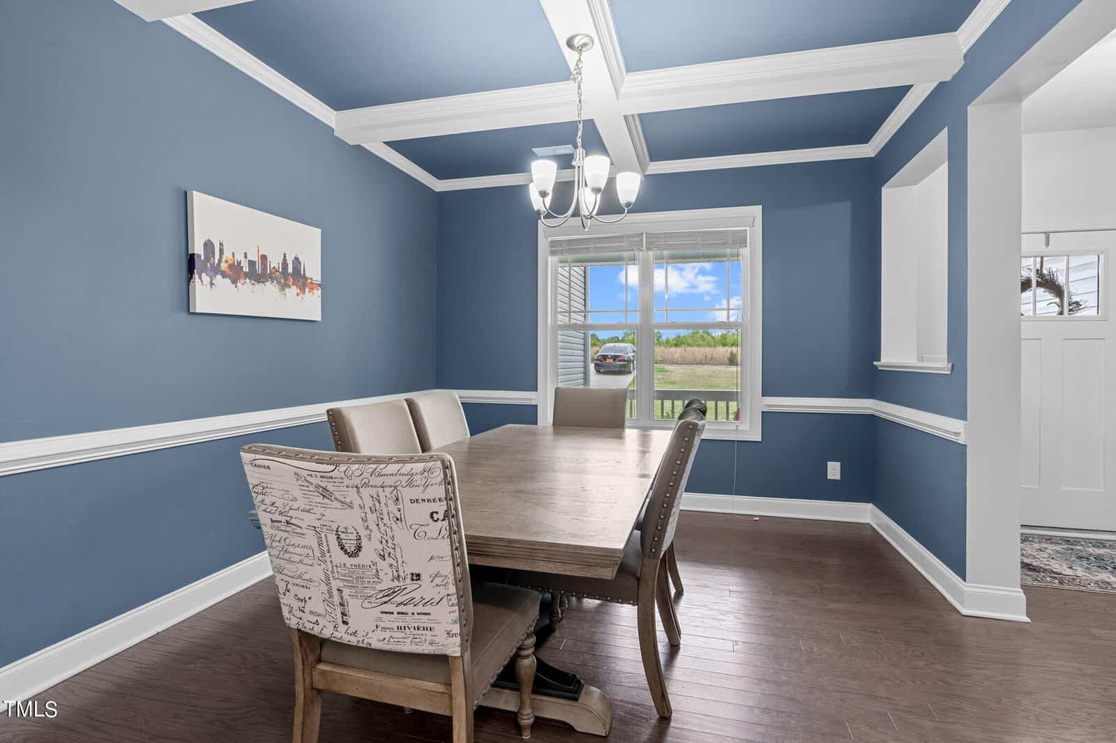 Dining room with blue walls and white trim, featuring a wooden table and chairs. One chair has printed upholstery. A chandelier hangs from a coffered ceiling, and there's a large window with a view of greenery. A cityscape painting adorns the wall.