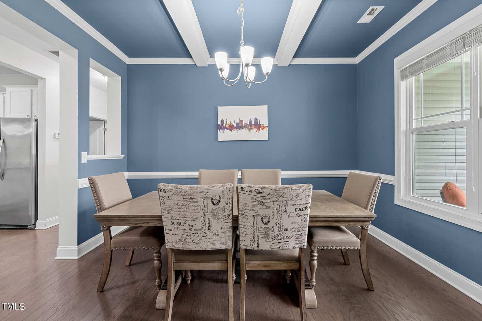 A dining room with a wooden table and six upholstered chairs, two of which have patterned fabric. The walls and ceiling are blue, with a chandelier hanging above. A small cityscape painting is on one wall, and a window is on the right.