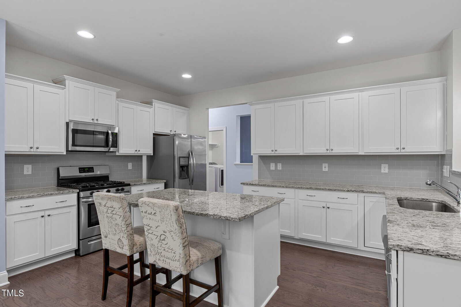 A modern kitchen with white cabinets and granite countertops. It features stainless steel appliances, including a refrigerator, oven, and microwave. An island with two patterned chairs stands in the center on a hardwood floor.