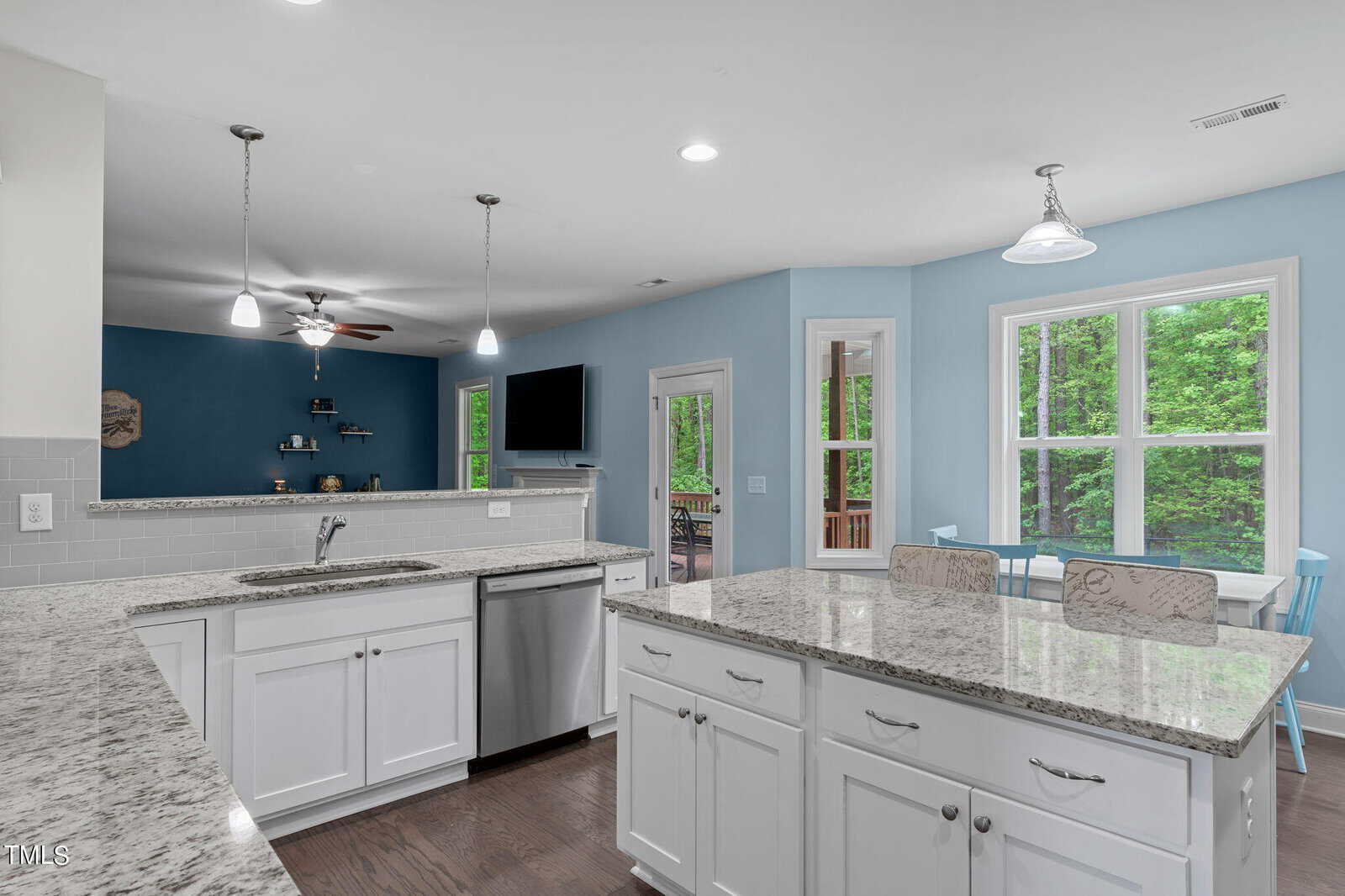 Modern kitchen with white cabinets, granite countertops, and stainless steel appliances. An island with seating faces a cozy living area with a blue accent wall and wall-mounted TV. Large windows and glass doors offer views of lush greenery outside.