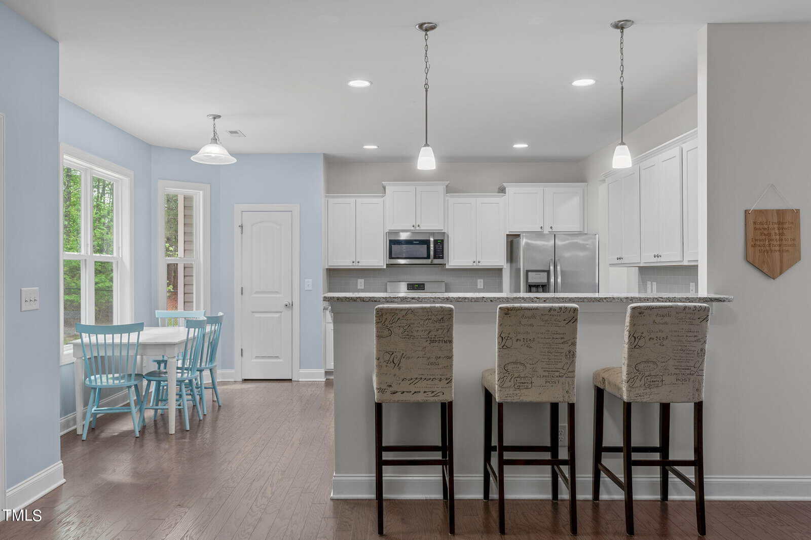Bright, modern kitchen with white cabinets and stainless steel appliances. A marble island with three upholstered bar stools faces the kitchen. A dining area with a round table and light blue chairs is near a window, illuminating the space.
