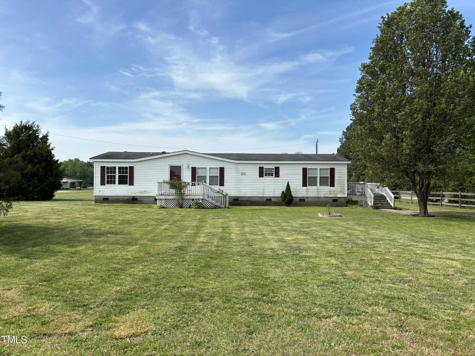 A white mobile home with a gray roof, featuring a front porch and stairs, positioned at 8340 Nc-39, Henderson NC on a large grassy lot with scattered trees and