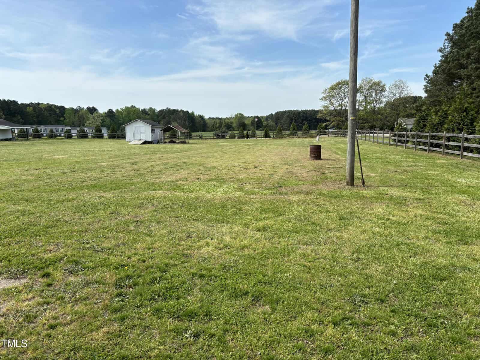 A wide open grassy field bordered by a wooden fence and trees with a small white shed in the distance, under a clear blue sky.